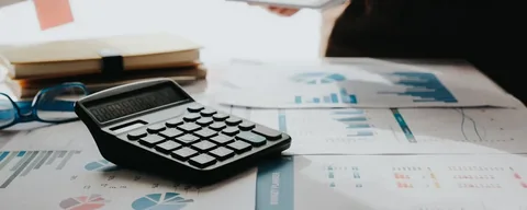 A calculator rests on top of documents and charts, representing the detailed financial analysis and due diligence involved in AI&GB's Project Management and Execution services for bid analysis and contract awarding.