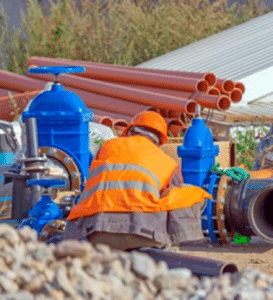 An engineering technician in safety gear at a job site with industrial pipes and valves, symbolizing the Strategic Procurement & Sourcing expertise offered by AI&GB Consulting.